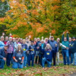 A group of people stand in front of a copse of trees on a bright autumn day.